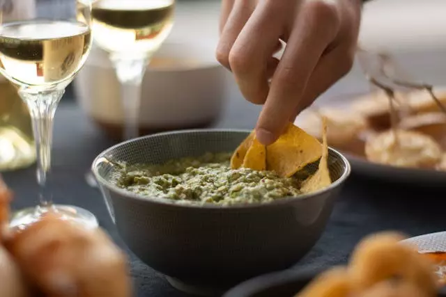 dłoń trzymająca nachos zanurzone w smacznie prezentującym się guacamole, obok z lewej strony stoją dwa kieliszki z białym winem.