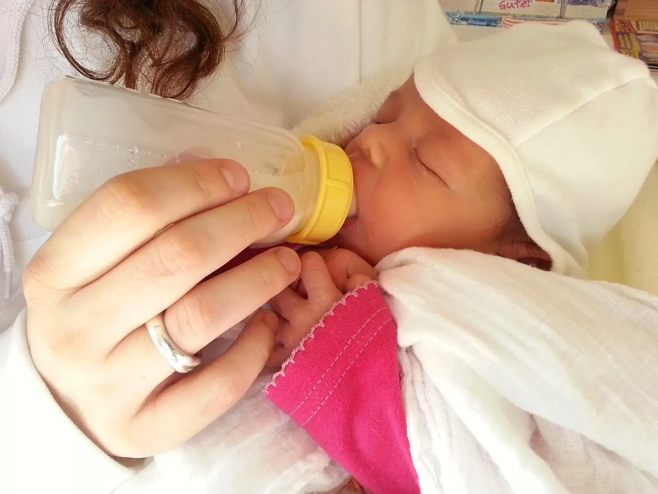 Mama karmiąca butelką noworodka w różowej koszulce i białej czapeczce.